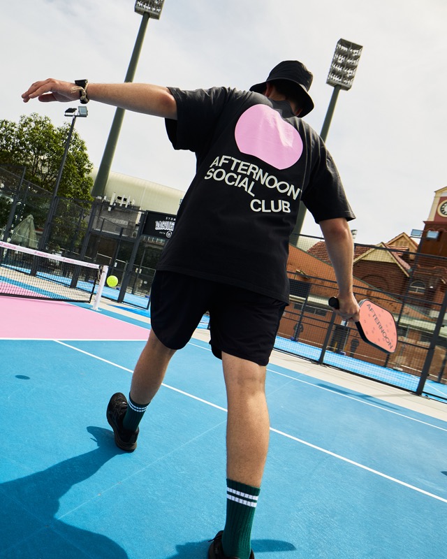 A player in an Afternoon Social Club tee mid-shot on a blue court